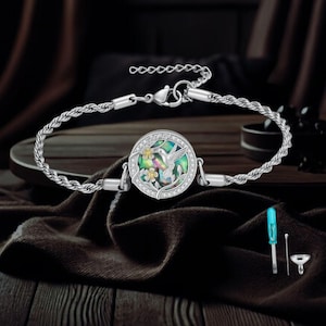 May include: Silver-toned bracelet featuring a round pendant with a hummingbird design. The pendant has a mother-of-pearl background with a hummingbird and floral accents. The bracelet includes a chain and a clasp. A small screwdriver and tool are included.