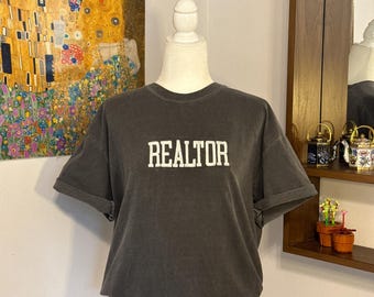 May include: A charcoal grey short-sleeved t-shirt featuring the word REALTOR printed in white block lettering across the chest. The garment has a crew neckline and cuffed sleeves, displayed on a white mannequin torso against a neutral background.