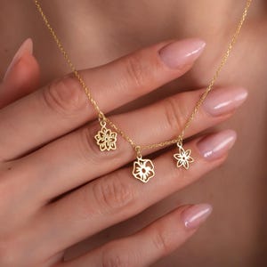Può includere: Una delicata collana d'oro con tre ciondoli a forma di fiore. I ciondoli presentano diversi disegni floreali, sospesi a una sottile catena d'oro. La collana è mostrata su uno sfondo color pelle.