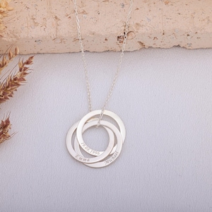 Op de afbeelding: Een zilveren ketting met drie in elkaar grijpende ringen. Elke ring is gegraveerd met een naam en een datum. De namen "Emma" en "Michael" zijn zichtbaar, samen met de datum "19.08.1996". De ketting hangt aan een fijne ketting.