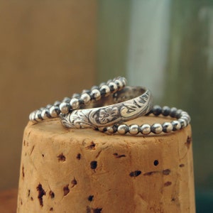 May include: Three silver rings stacked on top of each other. The top ring has a floral design, the middle ring is a simple band, and the bottom ring is a beaded band.