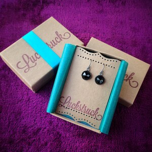 May include: Black gemstone earrings presented on a cardboard display card, with the word "Luckstruck" printed on it. The earrings are in a gift box with the word "Luckstruck" printed on it. The background is a purple fabric.