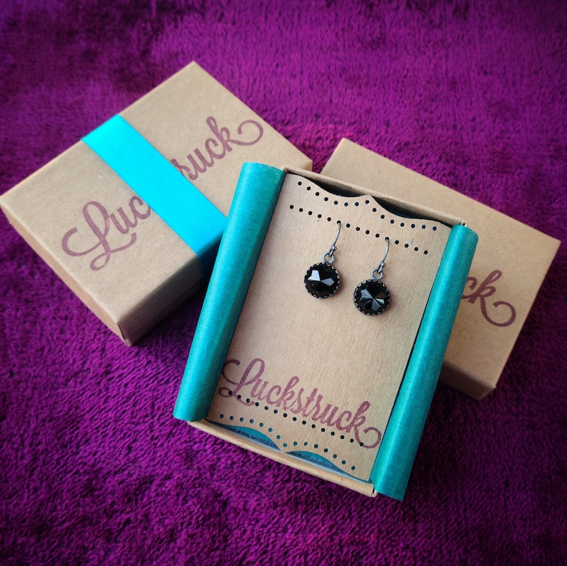 May include: Black gemstone earrings presented on a cardboard display card, with the word "Luckstruck" printed on it. The earrings are in a gift box with the word "Luckstruck" printed on it. The background is a purple fabric.