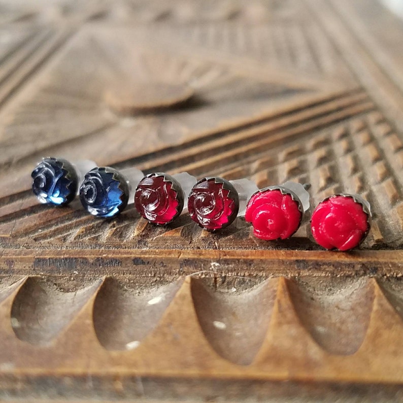 May include: Six rose-shaped stud earrings in shades of blue and red. The earrings are set on clear posts and feature a detailed floral design. They are displayed on a wooden surface with carved patterns.