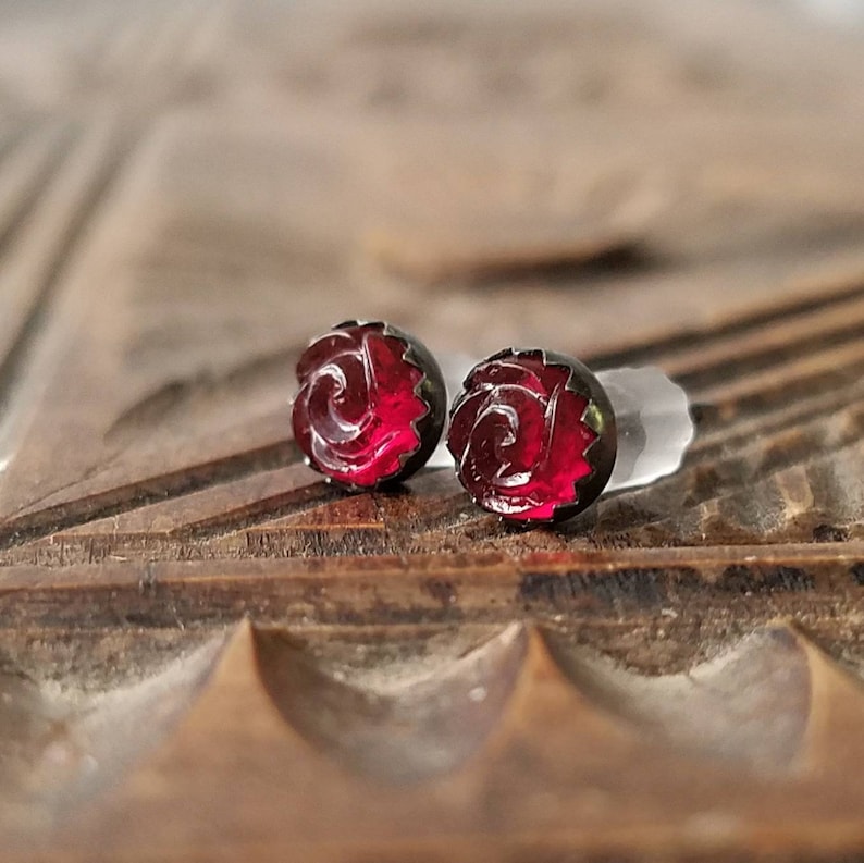 May include: A pair of stud earrings with a rose design. The earrings have a dark border and a vibrant red centre. The rose design is carved into the red centre. The earrings are displayed on a wooden surface with carved details.