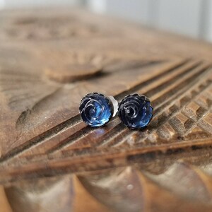 May include: A pair of dark blue rose-shaped stud earrings. The earrings are made of a translucent material, with intricate detailing that mimics the petals of a rose. The earrings are set on silver posts.