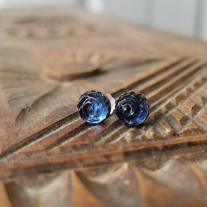 May include: A pair of dark blue rose-shaped stud earrings. The earrings are made of a translucent material, with intricate detailing that mimics the petals of a rose. The earrings are set on silver posts.