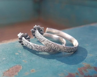 May include: A pair of silver hoop earrings with an intricate floral design. The earrings are made of sterling silver and have a polished finish.
