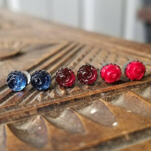 May include: Six rose-shaped stud earrings in different colours: two dark blue, two red, and two bright red. The earrings are displayed on a carved wooden surface, highlighting their floral design and vibrant hues.