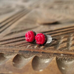 May include: A pair of red rose stud earrings with black trim. The earrings are small and detailed, featuring a classic floral design. The earrings are set against a wooden background.