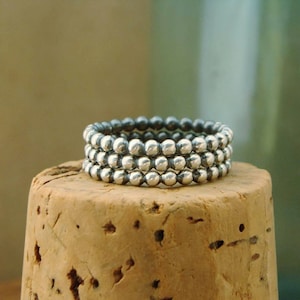 May include: Three silver beaded rings stacked on top of each other. The rings are made of small, round beads that are silver in color. The rings are on a cork surface.