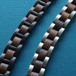 May include: Two wooden watch straps with a dark brown wood grain and metal links. One strap has dark grey metal links, and the other has silver metal links. The straps are on a textured blue background.