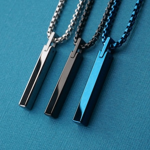 May include: Three metallic pendant necklaces in silver, gunmetal, and blue. Each necklace features a rectangular pendant suspended from a matching chain. The pendants are displayed against a textured blue background.