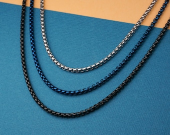 May include: Three round box chain necklaces in silver, blue, and black. The necklaces are displayed on a blue surface with a tan background. The chains are close together, showing the details of the links.
