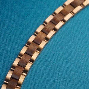 May include: A wooden bracelet with a gold-colored metal band. The bracelet features alternating dark brown wooden blocks and gold-colored metal links. The bracelet is curved and set against a blue background.