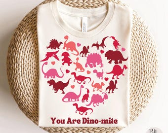 May include: Cream-colored t-shirt featuring a heart-shaped design of various dinosaurs in shades of pink and red. The phrase "You Are Dino-mite" is printed below the heart. The shirt is folded on a woven, circular surface.
