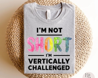 May include: A heather gray t-shirt featuring the black text I'M NOT, followed by the word SHORT in colorful watercolor letters, and the text I'M VERTICALLY CHALLENGED in black. The shirt has a crew neckline and a white brand label at the collar.