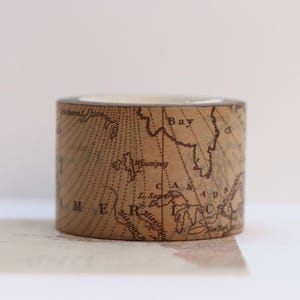 Puede incluir: Un rollo de cinta adhesiva decorativa con un diseño de mapa vintage. La cinta es de color marrón claro con una impresión de mapa detallada en marrón oscuro. El mapa incluye la palabra "AMERICA" y otros detalles geográficos.