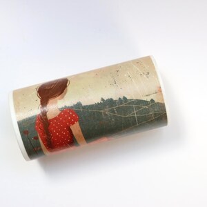 May include: A white ceramic mug with a colorful illustration of a woman with long brown hair wearing a red dress with white polka dots. The woman is standing in a field with green trees and a blue sky with white clouds. The mug is wrapped in a brown paper with a white string.