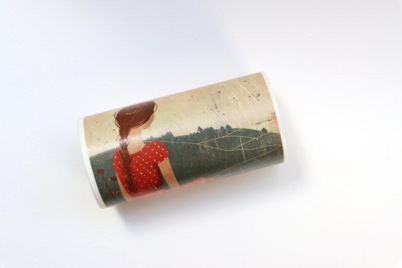 May include: A white ceramic mug with a colorful illustration of a woman with long brown hair wearing a red dress with white polka dots. The woman is standing in a field with green trees and a blue sky with white clouds. The mug is wrapped in a brown paper with a white string.