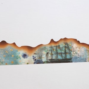Può includere: Una striscia di carta decorativa con un collage di elementi in stile vintage, tra cui un veliero scuro, scrittura a mano e motivi floreali blu. Il bordo superiore della carta presenta un bordo marrone bruciato e irregolare.