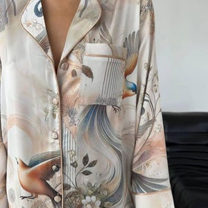 Könnte beinhalten: Cremefarbenes Pyjama-Set mit Vogel- und Blumendruck. Das langärmlige Oberteil hat einen Reverskragen, eine Brusttasche und Knopfverschlüsse. Das Design umfasst Vögel mit blauen, orangefarbenen und braunen Federn sowie florale und botanische Elemente.