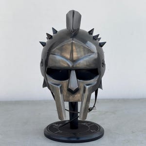 Könnte beinhalten: Ein silberner Gladiatorenhelm mit schwarzem Visier und Spikes auf der Oberseite. Der Helm steht auf einem schwarzen Ständer.