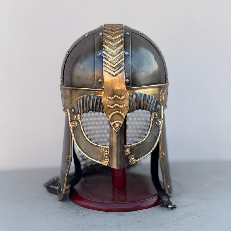 May include: A silver and gold Viking helmet with a chainmail face mask and a decorative crest. The helmet is on a red wooden stand.
