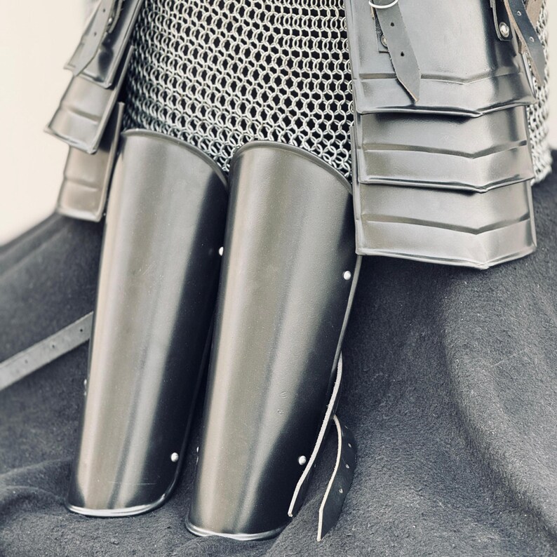 May include: A pair of black leather shin guards with metal accents. The shin guards are worn over chainmail armor.