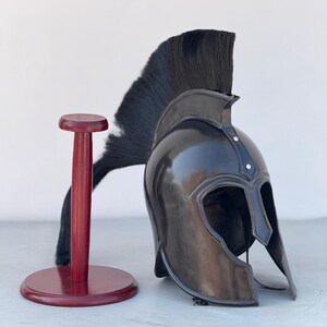May include: A black Spartan helmet with a black horsehair plume, displayed on a wooden stand. The helmet has a distinctive crest and cheek guards.