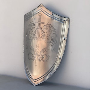 May include: A silver shield with a sword and two dragons etched into the surface. The shield has a rounded top and a pointed bottom.