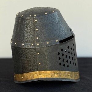 May include: A black metal helmet with a gold trim and a decorative pattern. The helmet has a visor with a diamond-shaped pattern.