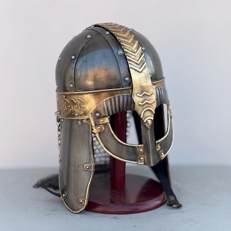 May include: A Viking helmet with a gold trim and a chainmail face mask. The helmet is on a wooden stand.