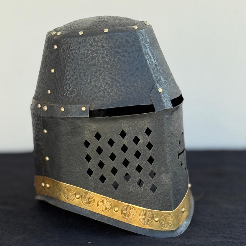 May include: A black metal helmet with a gold trim and a cross design. The helmet has a face shield with a diamond pattern.