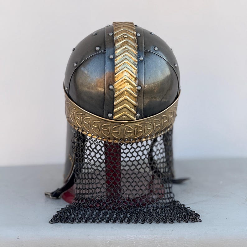 May include: A black and gold Viking helmet with a chainmail face mask. The helmet has a decorative band with a Celtic knot design and a gold chevron pattern on the top.