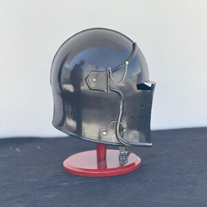 May include: A black helmet with a visor and a cross on the side. The helmet is on a wooden stand.