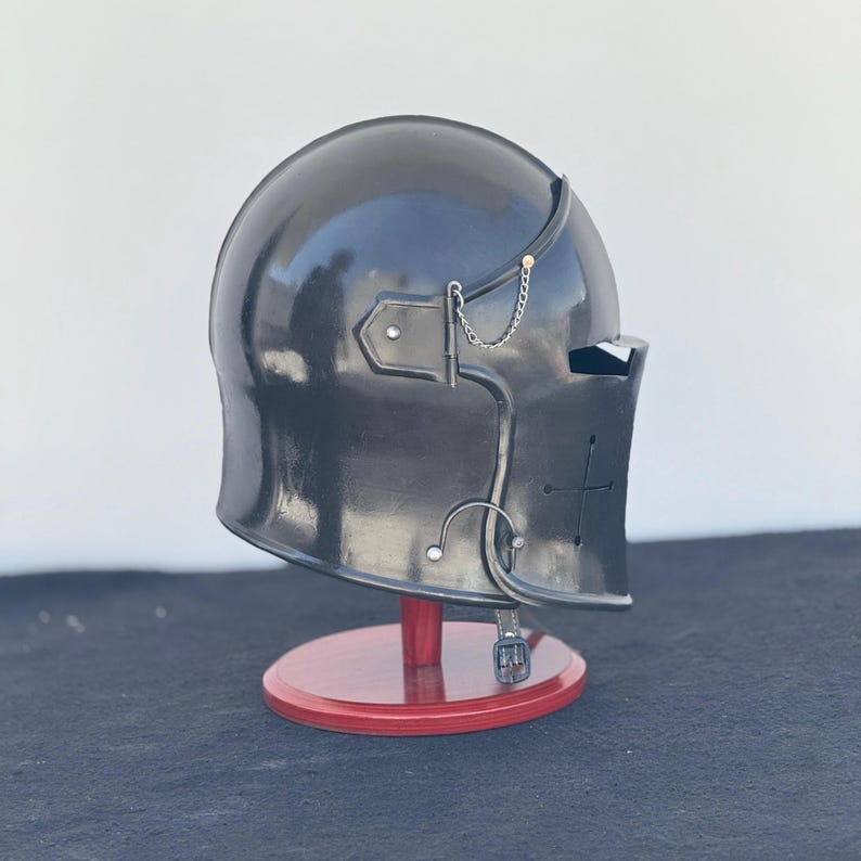 May include: A black helmet with a visor and a cross on the side. The helmet is on a wooden stand.
