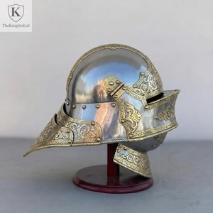 Op de afbeelding: Een gepolijste stalen helm met sierlijke goudkleurige filigraan en accenten. De helm heeft een puntig vizier en wordt tentoongesteld op een houten standaard. Het logo TheKnightsLtd staat in de linkerbovenhoek.
