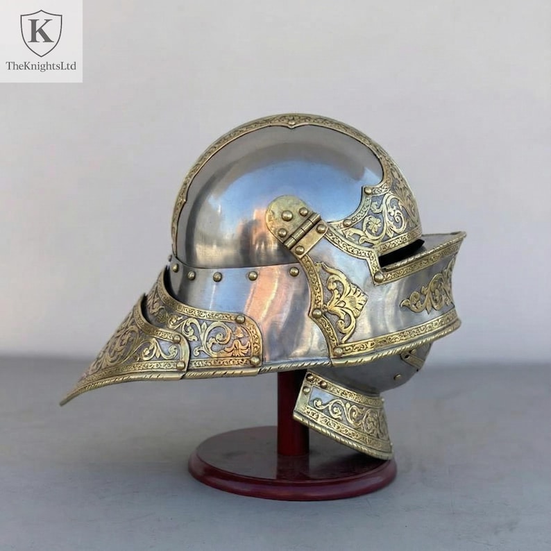 Op de afbeelding: Een gepolijste stalen helm met sierlijke goudkleurige filigraan en accenten. De helm heeft een puntig vizier en wordt tentoongesteld op een houten standaard. Het logo TheKnightsLtd staat in de linkerbovenhoek.