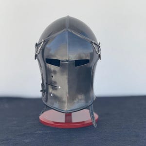 May include: A black metal helmet with a visor and chainmail accents. The helmet is on a red display stand.