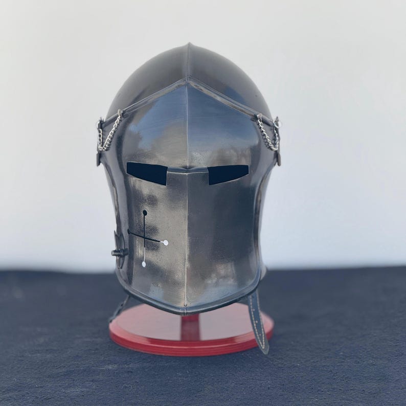 May include: A black metal helmet with a visor and chainmail accents. The helmet is on a red display stand.