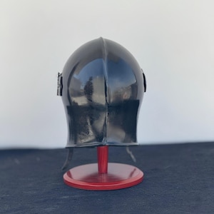 May include: A black helmet with a pointed top and a visor. The helmet is on a red wooden stand.