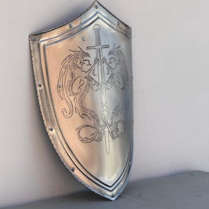 May include: A silver shield with a sword and two dragons etched into the surface. The shield is a traditional shape with a pointed bottom.