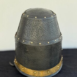 May include: A black helmet with a textured surface and gold accents. The helmet has a band of gold around the bottom with a repeating pattern of four-leaf clover designs.