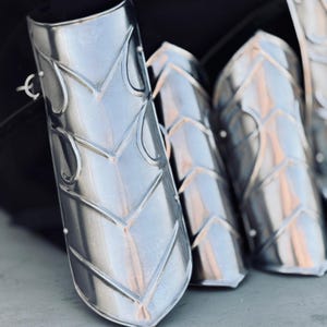 K&ouml;nnte beinhalten: Nahaufnahme von vier silbernen Armr&uuml;stungen aus Metall mit einem dekorativen Muster. Die R&uuml;stungen sind aus Metall gefertigt und haben eine gl&auml;nzende Oberfl&auml;che.