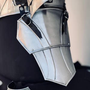 K&ouml;nnte beinhalten: Silbernes Metall-R&uuml;stungsteil mit spitzem Design und schwarzen Lederriemen. Die R&uuml;stung besteht aus mehreren Platten und hat eine glatte, polierte Oberfl&auml;che.