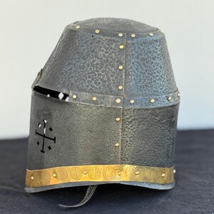 May include: A black metal helmet with a textured surface and gold accents. The helmet has a cross design on the side and a band of gold with circular designs around the bottom.