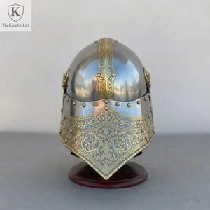 Op de afbeelding: Een gepolijste zilveren helm met sierlijke goudkleurige accenten. De helm heeft ingewikkelde bloempatronen en wordt gepresenteerd op een houten standaard. Het logo van The Knights Ltd staat linksboven.