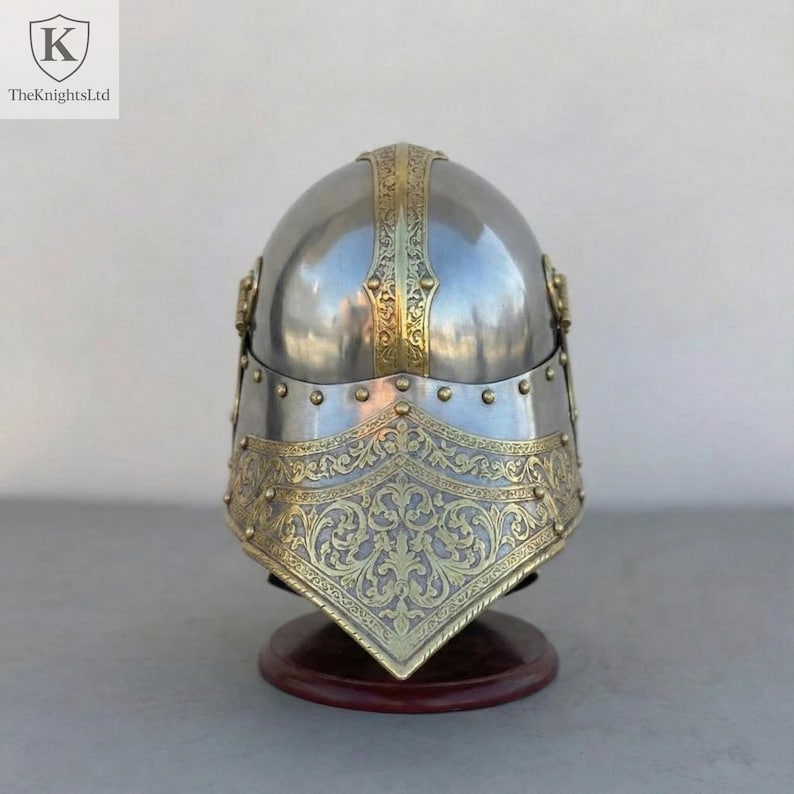 Op de afbeelding: Een gepolijste zilveren helm met sierlijke goudkleurige accenten. De helm heeft ingewikkelde bloempatronen en wordt gepresenteerd op een houten standaard. Het logo van The Knights Ltd staat linksboven.