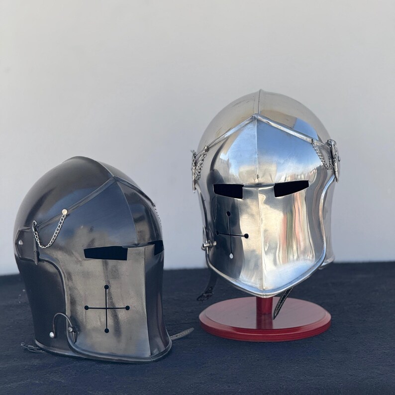 May include: Two medieval helmets, one black and one silver, with a cross design on the faceplate. Both helmets are on display stands.